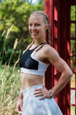 Portrait of smiling fit young caucasian woman leaning against an old classic red phone box. She is wearing a white tennis skirt and sport bra. She is in a park.
