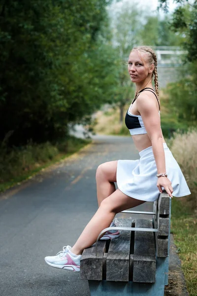公園のベンチに座っている若い白人女性の肖像画 彼女は白いテニススカートとスポーツブラジャーを着ています — ストック写真