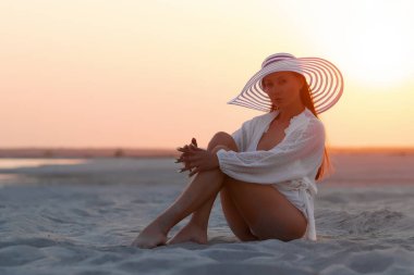 Seksi gömlekli ve şapkalı çekici kadın gün batımında kumlu sahilde şehvetle oturuyor.