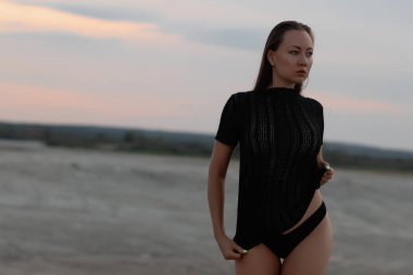 Alacakaranlıkta kumlu sahilde duran siyah şeffaf tişörtlü, seksi külotlu genç bir esmer.