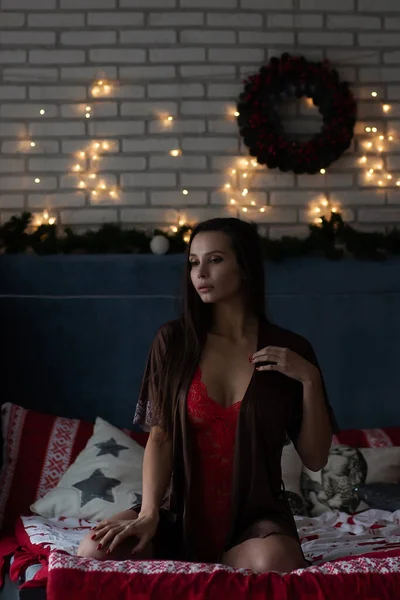 自己保証若いです女性で茶色の絹のローブと赤いボディスーツに座って快適なベッドで装飾された居心地の良い部屋で輝くガーランドとクリスマス休暇中の花輪 — ストック写真