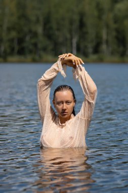 Yumuşak, baştan çıkarıcı, beyaz, ıslak, şeffaf gömlekli, gölette duran ve başka tarafa bakan bir kadın.