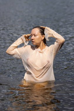 Yumuşak, baştan çıkarıcı, beyaz, ıslak, şeffaf gömlekli, gölette duran ve başka tarafa bakan bir kadın.