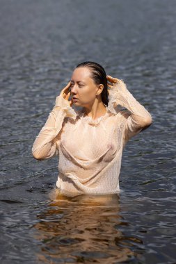 Yumuşak, baştan çıkarıcı, beyaz, ıslak, şeffaf gömlekli, gölette duran ve başka tarafa bakan bir kadın.