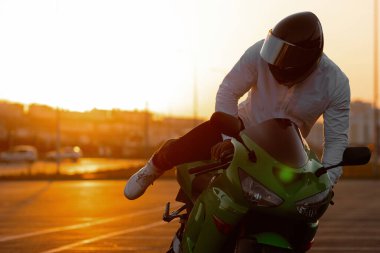 Tanımlanamayan stil sahibi, kasklı erkek motosikletçi, günbatımı gökyüzünün arka planında, park halindeki motosiklette oturuyor.
