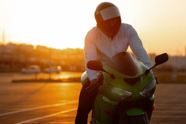 Tanımlanamayan stil sahibi, kasklı erkek motosikletçi, günbatımı gökyüzünün arka planında, park halindeki motosiklette oturuyor.