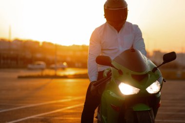 Tanımlanamayan stil sahibi, kasklı erkek motosikletçi, günbatımı gökyüzünün arka planında, park halindeki motosiklette oturuyor.