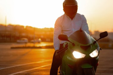 Tanımlanamayan stil sahibi, kasklı erkek motosikletçi, günbatımı gökyüzünün arka planında, park halindeki motosiklette oturuyor.