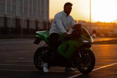 Yakışıklı erkek motosikletlinin, asfalt park yerinde günbatımına karşı park etmiş modern havalı motosikletinin üzerinde oturuşunun yan görüntüsü