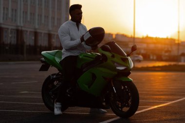 Yakışıklı erkek motosikletlinin, asfalt park yerinde günbatımına karşı park etmiş modern havalı motosikletinin üzerinde oturuşunun yan görüntüsü