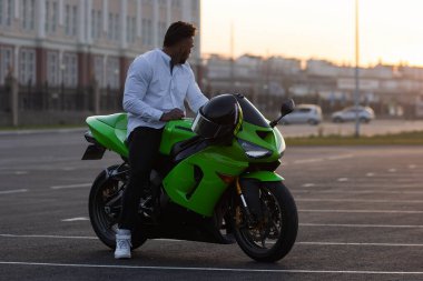 Yakışıklı erkek motosikletlinin, asfalt park yerinde günbatımına karşı park etmiş modern havalı motosikletinin üzerinde oturuşunun yan görüntüsü