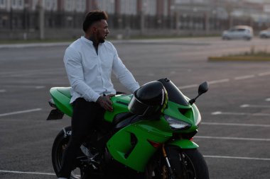 Yakışıklı erkek motosikletlinin, asfalt park yerinde günbatımına karşı park etmiş modern havalı motosikletinin üzerinde oturuşunun yan görüntüsü