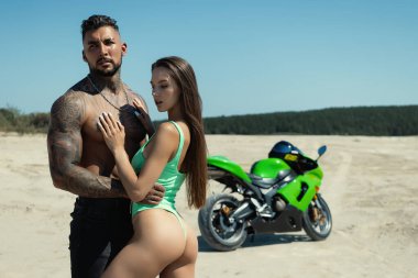 Ciddi bir erkek, sakallı, ayakta duran, uzun kahverengi saçlı, güneşli bir günde kumlu arazide yeşil motorsiklete karşı şehvetli bir kız arkadaşı olan.