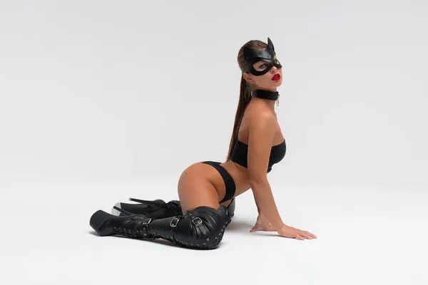 Full body side view of alluring slim tanned brunette in black mask and underwear with high heeled leather boots in white studio