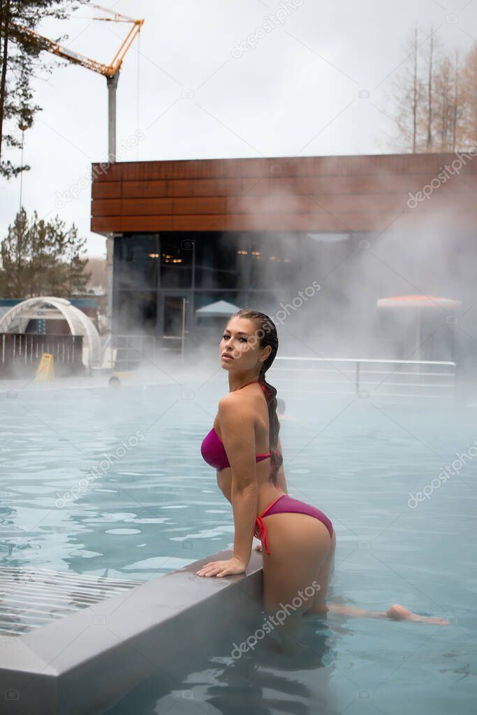 Side view of sensual slim female in red bikini leaning on border and looking at camera in warm water of hot spring