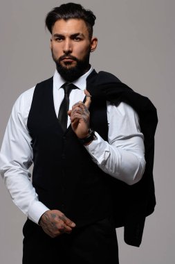Bearded Hispanic guy in dark vest and white shirt with tie looking at camera with hands in pockets in studio clipart