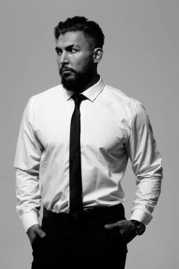 Siyah yelekli, beyaz gömlekli, sakallı adam stüdyoda elleri ceplerinde kameraya bakıyor.