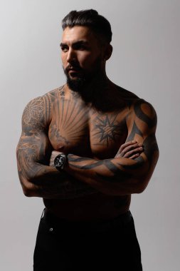 Hispanic shirtless male model with muscular tattooed torso standing with hands in pockets and looking away on gray backdrop clipart