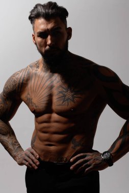 Hispanic shirtless male model with muscular tattooed torso standing with hands in pockets and looking away on gray backdrop clipart