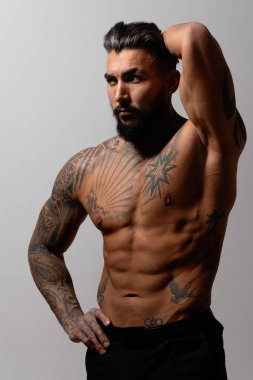 Kaslı gövdesi dövmeli İspanyol tişörtsüz erkek manken elleri ceplerinde duruyor ve gri arka plana bakıyor.