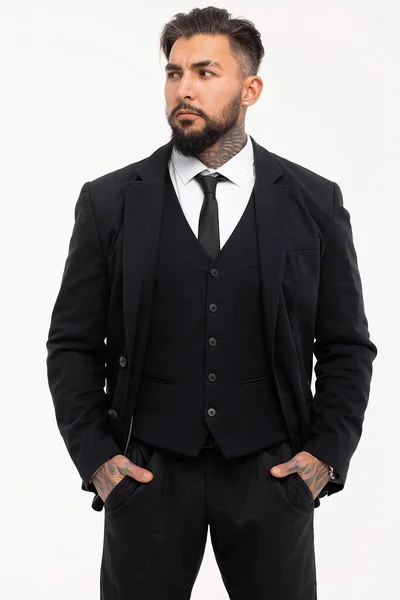 Siyah yelekli, beyaz gömlekli, sakallı adam stüdyoda elleri ceplerinde kameraya bakıyor.