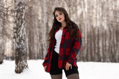 Karlı kış ormanlarında dikilip kameraya bakarken kırmızı dudaklı, karlı gömlekli, saçlarına dokunan çekici bir kadın.