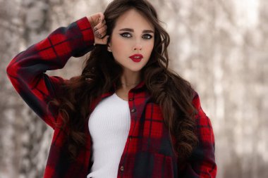 Karlı kış ormanlarında dikilip kameraya bakarken kırmızı dudaklı, karlı gömlekli, saçlarına dokunan çekici bir kadın.