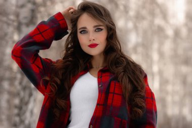 Karlı kış ormanlarında dikilip kameraya bakarken kırmızı dudaklı, karlı gömlekli, saçlarına dokunan çekici bir kadın.