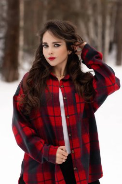 Karlı kış ormanlarında dikilip kameraya bakarken kırmızı dudaklı, karlı gömlekli, saçlarına dokunan çekici bir kadın.