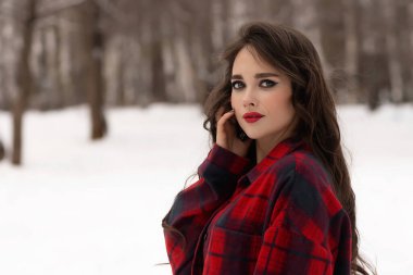 Karlı kış ormanlarında dikilip kameraya bakarken kırmızı dudaklı, karlı gömlekli, saçlarına dokunan çekici bir kadın.