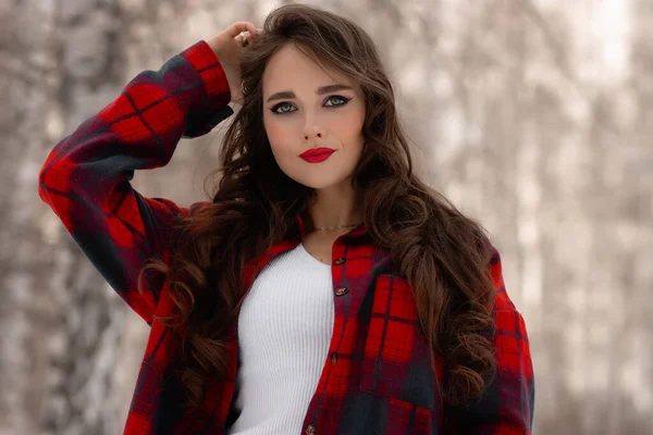 Karlı kış ormanlarında dikilip kameraya bakarken kırmızı dudaklı, karlı gömlekli, saçlarına dokunan çekici bir kadın.