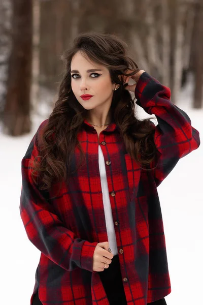 Karlı kış ormanlarında dikilip kameraya bakarken kırmızı dudaklı, karlı gömlekli, saçlarına dokunan çekici bir kadın.