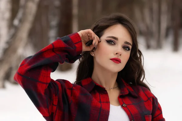 雪の冬の森の中に立ち カメラを見ている間 赤い唇とチェックされたシャツで魅力的な女性 — ストック写真