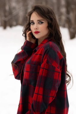 Karlı kış ormanlarında dikilip kameraya bakarken kırmızı dudaklı, karlı gömlekli, saçlarına dokunan çekici bir kadın.