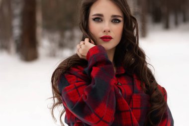 Karlı kış ormanlarında dikilip kameraya bakarken kırmızı dudaklı, karlı gömlekli, saçlarına dokunan çekici bir kadın.