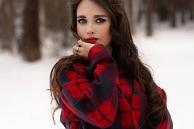 Karlı kış ormanlarında dikilip kameraya bakarken kırmızı dudaklı, karlı gömlekli, saçlarına dokunan çekici bir kadın.