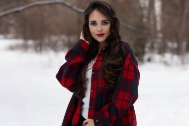 Karlı kış ormanlarında dikilip kameraya bakarken kırmızı dudaklı, karlı gömlekli, saçlarına dokunan çekici bir kadın.