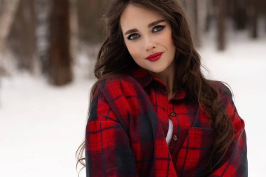 Karlı kış ormanlarında dikilip kameraya bakarken kırmızı dudaklı, karlı gömlekli, saçlarına dokunan çekici bir kadın.