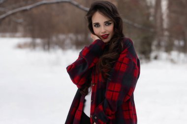 Karlı kış ormanlarında dikilip kameraya bakarken kırmızı dudaklı, karlı gömlekli, saçlarına dokunan çekici bir kadın.