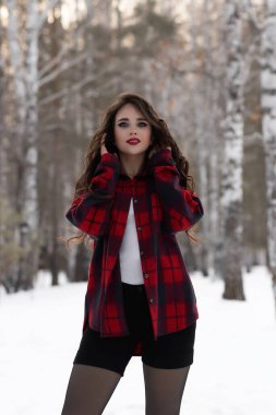 Karlı kış ormanlarında dikilip kameraya bakarken kırmızı dudaklı, karlı gömlekli, saçlarına dokunan çekici bir kadın.