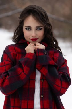 Karlı kış ormanlarında dikilip kameraya bakarken kırmızı dudaklı, karlı gömlekli, saçlarına dokunan çekici bir kadın.