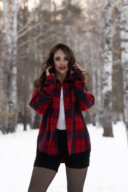 Karlı kış ormanlarında dikilip kameraya bakarken kırmızı dudaklı, karlı gömlekli, saçlarına dokunan çekici bir kadın.