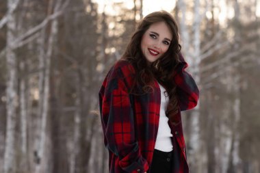 Karlı kış ormanlarında dikilip kameraya bakarken kırmızı dudaklı, karlı gömlekli, saçlarına dokunan çekici bir kadın.