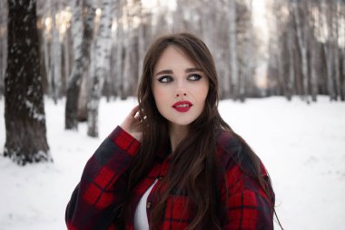 Karlı kış ormanlarında dikilip kameraya bakarken kırmızı dudaklı, karlı gömlekli, saçlarına dokunan çekici bir kadın.