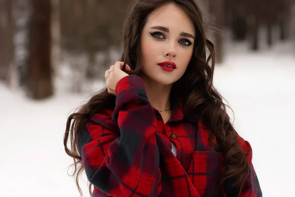 雪の冬の森の中に立ち カメラを見ている間 赤い唇とチェックされたシャツで魅力的な女性 — ストック写真