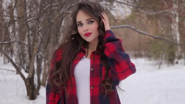 Kış ormanında dalgalı saçlı genç bir kadın ayakta duruyor ve yüzüne dokunuyor. — Stok video