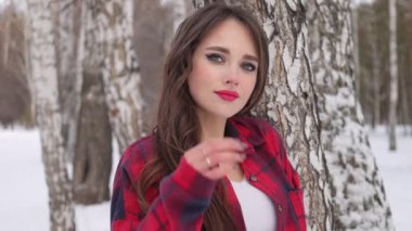 Kış ormanında dalgalı saçlı genç bir kadın ayakta duruyor ve yüzüne dokunuyor.