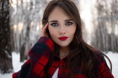 Karlı kış ormanlarında dikilip kameraya bakarken kırmızı dudaklı, karlı gömlekli, saçlarına dokunan çekici bir kadın.