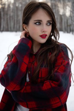 Karlı kış ormanlarında dikilip kameraya bakarken kırmızı dudaklı, karlı gömlekli, saçlarına dokunan çekici bir kadın.