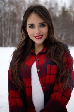 Karlı kış ormanlarında dikilip kameraya bakarken kırmızı dudaklı, karlı gömlekli, saçlarına dokunan çekici bir kadın.